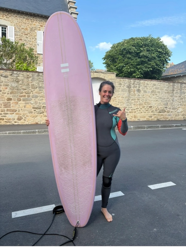 Omblyne en combinaison, debout à côté de son longboard, à Saint-Malo